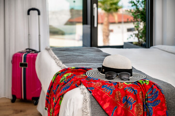 Featuring a summer brimmed hat and sunglasses arranged on the edge of a bed, complemented by a colorful pareo. In the blurred background, a suitcase adds to the anticipation of a journey.