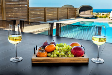 Summer Soiree: Close-Up of Two White Wine Glasses Flanked by a Tray of Fresh Fruits, Poolside Bliss in the Background. Perfect for conveying the essence of a leisurely summer gathering.