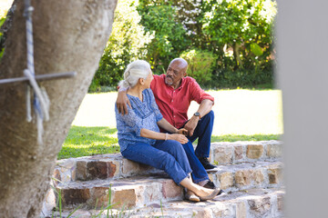 Happy diverse senior couple embracing and sitting on stairs in sunny garden