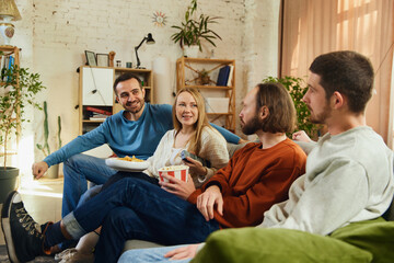 Group portrait. Guys and girl sitting on couch in living room and discussing while watching comedy...