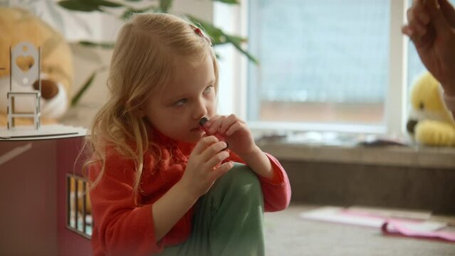 Cute toddler girl in process of applying lipbalm on her lips, smelling it's aroma, side view, beauty care, girl's growing. Female beauty routine, sunny nursery room background. High quality 4k footage
