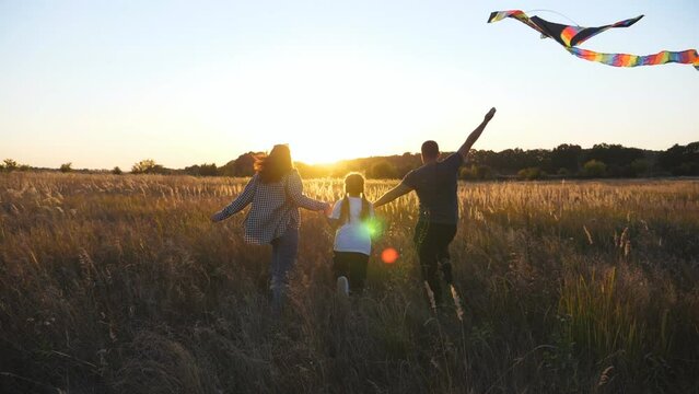 Young parents with small daughter running with multicolored kite through grass field at sunset. Mother and father with little cute girl holds hands of each other jogging among summer meadow. Rear view