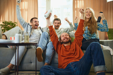 Bottom view of cheerful group of friends sitting on sofa in living room and watching basketball match and favorite team win victory, - obrazy, fototapety, plakaty