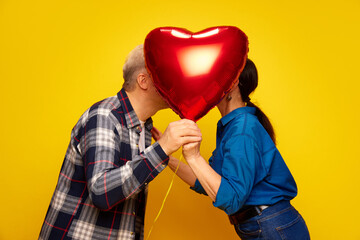 Senior couple, wife and husband standing behind red balloon in heart shape and kissing against yellow studio background. Concept of marriage, relationship, Valentine's Day, love, emotions, fashion