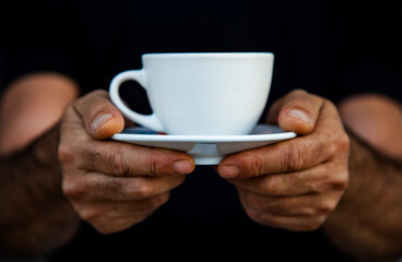 man's hands holding a cup of coffee. Close-up of white mug on saucer against black background. 
