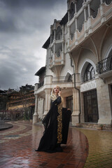 A female queen, blonde in a black dress, sits on a wooden throne in a concrete room next to candlesticks