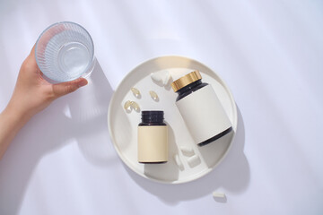 Two unlabeled bottles of dietary supplements are displayed on a white ceramic tray. A hand holds a glass of water next to it. Minimalist white background.