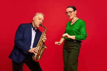 Naklejka premium Caring, loving man, husbands playing saxophone to his beautiful wife against red studio background. Romantic date, holiday. Concept of marriage, relationship, Valentine's Day, love, emotions, fashion