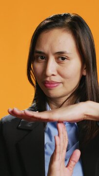Vertical Video Serious adult presenting timeout gesture in studio, asking for a break after working too hard at the office. Asian woman standing over orange background, doing pause symbol with hands.
