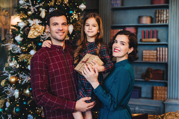 Obraz na płótnie Canvas Cheerful family with their daughter holding a Christmas gift, standing in front of a tree in a festively decorated room