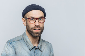 Surprised man with a beard in glasses and a beanie, wearing a denim shirt, against a grey background - obrazy, fototapety, plakaty