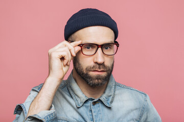 Obraz premium Serious man adjusting glasses, wearing beanie and denim shirt, against a pink background