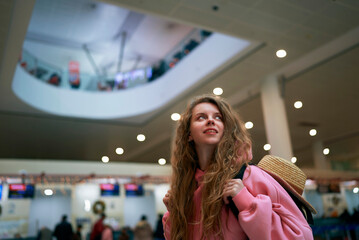 Smiling woman in pink jacket carries straw hat, strolling in bustling airport adorned with festive lights, holiday traveler embarks on a journey.