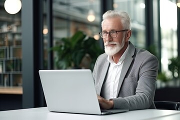 Old businessman sitting in front of a laptop in a modern office, coworking space. A gray-haired serious man works alone.