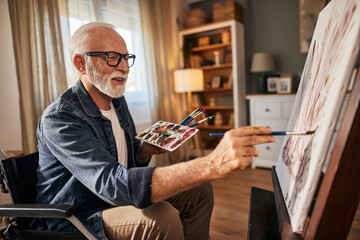 Senior man in a wheelchair painting on a canvas at home