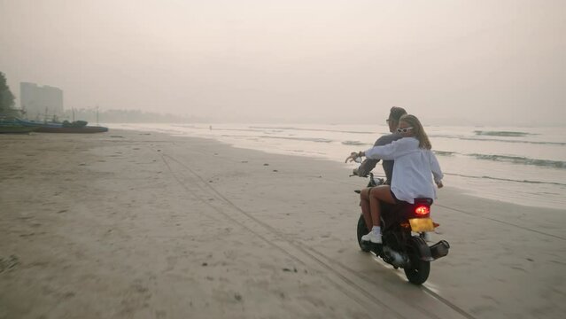 Young folks ride motorbike on sea beach on tropical island at sunset. Man, woman travel by scooter on coastline along ocean. Lady hugs boyfriend, spreads hands on moped adventure. Motivational video.