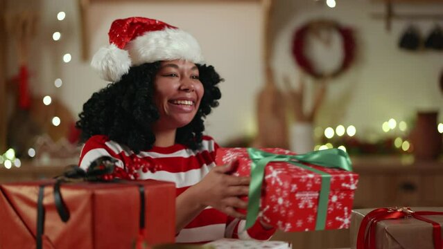 Smiling middle-aged black woman sitting in a cozy festively decorated room, wrapping presents for Christmas, holding out the box in red wrapping paper. High quality 4k footage