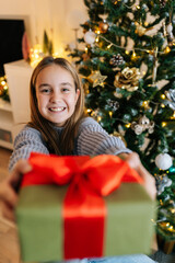 Vertical portrait of cheerful cute little girl holding, giving and showing at camera wrapped with red ribbon and bow festive Christmas present gift box, sitting by glowing xmas tree, looking at camera