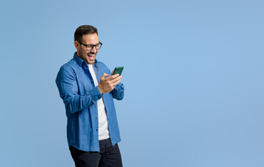 Young businessman laughing ecstatically while reading messages over mobile phone on blue background - obrazy, fototapety, plakaty