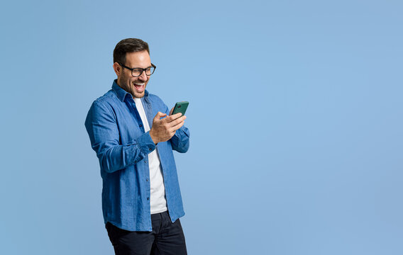 Young businessman laughing ecstatically while reading messages over mobile phone on blue background