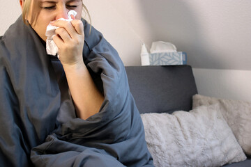 Sick woman sitting on sofa with blanket and sneezing in a tissue
