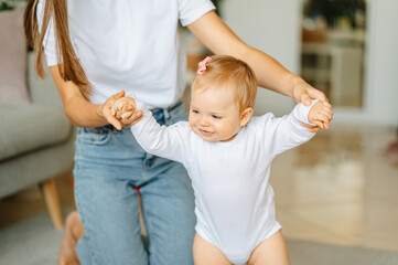 Cheerful baby girl is making the first steps walking with her moms help at home.