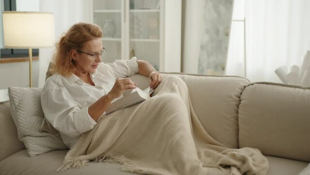 Pretty neat lady in glasses sitting on cozy sofa covering legs with plaid in a light living-room with modern renovation, turning pages of thick book. High quality 4k footage