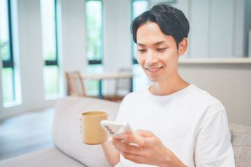 ソファに座ってコーヒーを飲みながらスマートフォンを操作する男性