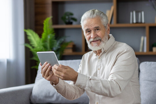 Close-up Portrait Of An Older Gray-haired Man Sitting On The Couch At Home, Holding A Tablet And Smiling At The Camera