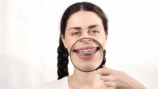 Portrait of young Caucasian smiling woman showing x-ray, magnifier glass and teeth with ligature braces. White background. Concept of orthodontic treatment