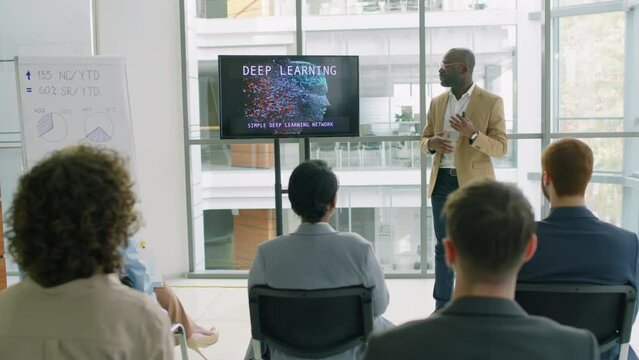 Panning shot of African American public speaker standing in light conference room showing presentation in front of his colleagues