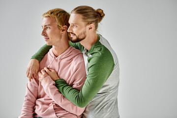 romantic and stylish gay couple embracing and looking away while standing on grey backdrop