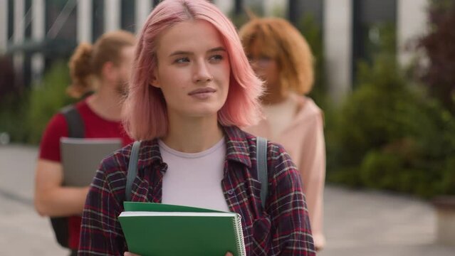 Caucasian alone lonely student girl with pink hair walking smiling at camera holding book walk in city outdoors multiracial diverse student university friends bulling college friendship socialization
