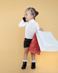 Cute little girl holding a package in hand and talking on a phone, dressed in a casual clothes, posing in studio, isolated beige background.