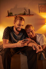 smiling bearded gay man browsing internet on mobile phone near happy boyfriend in bedroom at night