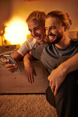 joyful bearded gay man browsing internet on mobile phone near smiling boyfriend in bedroom at night