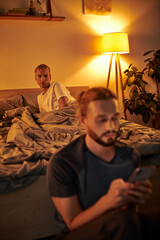 discouraged gay man looking at boyfriend messaging on mobile phone in bedroom at night, cheating
