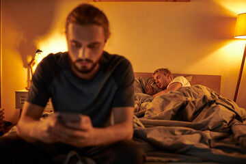 disloyal bearded gay man browsing internet on smartphone near partner sleeping at night in bedroom