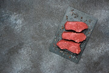 Three raw beef steaks on a black slate board on a dark concrete background. Steak ingredients, raw...
