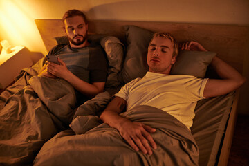 unfaithful bearded gay messaging on mobile phone near sleeping boyfriend at night in bedroom - obrazy, fototapety, plakaty