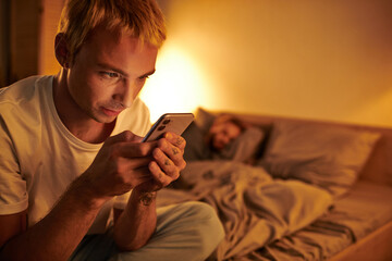 disloyal gay man messaging on mobile phone near boyfriend sleeping at night in bedroom, cheating