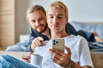 tattooed gay man with coffee cup browsing internet on mobile phone near smiling boyfriend in bedroom