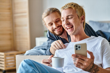 cheerful tattooed gay man with coffee cup browsing internet on smartphone near smiling boyfriend