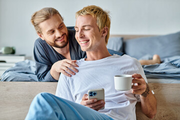 cheerful bearded gay man smiling near boyfriend with coffee cup using mobile phone in bedroom