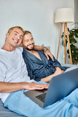 delighted gay couple sitting with closed eyes while watching romantic movie on laptop in bedroom