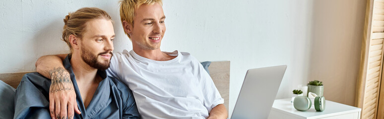 happy tattooed gay man embracing bearded boyfriend while watching film on laptop in bedroom, banner