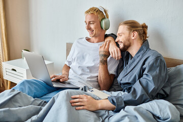 cheerful gay man in headphones using laptop near bearded boyfriend with smartphone in bedroom
