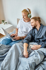 cheerful gay man in headphones using laptop near bearded boyfriend with smartphone in bedroom