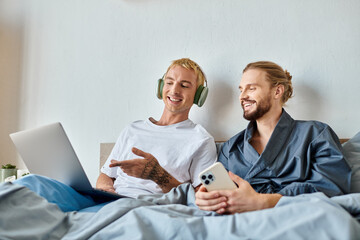 happy gay man in headphones pointing at laptop near bearded boyfriend with smartphone in bedroom