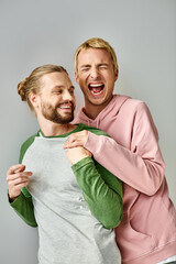 overjoyed and trendy gay man laughing with closed eyes near bearded boyfriend on grey backdrop